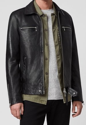 Homme portant une veste en cuir noire par-dessus une chemise olive boutonnée et un t-shirt gris clair avec un pantalon noir.