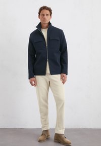 Marineblauwe zip-up jas met twee borstzakken, gecombineerd met beige broeken. De jas heeft een hoge kraag en een minimalistisch ontwerp.