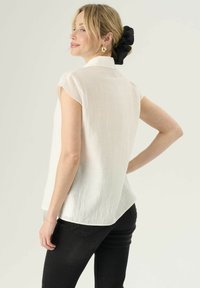 Blusa senza maniche in lino bianco con colletto, tessuto texturizzato, orlo curvo, indossata con jeans neri, con uno scrunchie nero nei capelli.