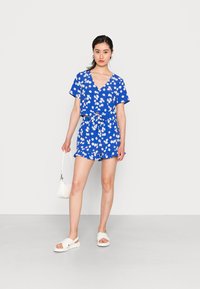 Macacão azul floral com mangas curtas, decote em V e cintura cingida; apresenta padrões de margaridas brancas e bainha com babados; combinado com sandálias brancas.