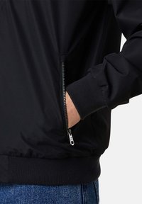 Giacca bomber nera con tasca con zip, polsini a coste e tessuto liscio e texturizzato. Cuciture visibili e vita elastica.