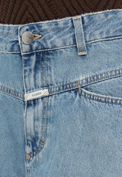Φωτεινό μπλε τζιν με ξεθωριασμένο φινίρισμα, διαθέτει κλείσιμο με κουμπί και ετικέτα με την επιγραφή "CLOSED." Ορατές ραφές και τσέπες.
