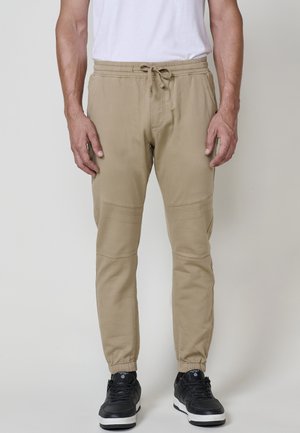 Hombre vestido con pantalones jogger beige con cordón y zapatillas negras, de pie frente a un fondo claro y simple.