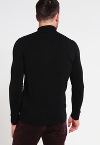 Homme tourné de dos portant un pull noir ajusté à manches longues et un pantalon bordeaux foncé sur un fond clair uni.
