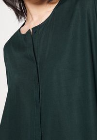 Camicia leggera di colore verde scuro con scollatura rotonda, chiusura a bottoni e una texture liscia con cuciture a contrasto sottili lungo la patta.