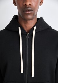 Sudadera con cremallera negra hecha de un tejido suave, que cuenta con una cremallera frontal y cordones color crema. Capucha ajustada y diseño minimalista.
