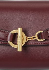 Detailný záber na bordovú koženú tašku so zlatým preklápacím uzáverom a reťazovou dekoráciou na prednej klapke.