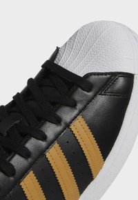 Black sneaker featuring three zigzag yellow stripes, black laces, textured white rubber toe cap, and a smooth leather upper.