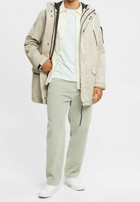 Beige vandtæt jakke med snoretræk i hætten over en hvid stribet skjorte, parret med lys grøn kordfløjl bukser og hvide sneakers.