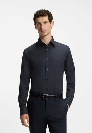 Homme portant une chemise habillée bleu marine foncé ajustée avec manches longues, boutonnée sur le devant, ceinture noire et pantalon foncé, debout avec une main dans la poche.