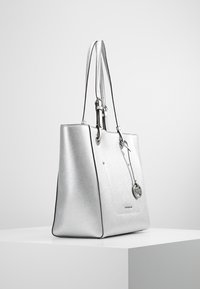 Srebrna torba tote z gładką metaliczną teksturą, podwójnymi długimi uchwytami i dekoracyjnym charmsiem. Charakteryzuje się eleganckim, ustrukturyzowanym designem oraz kontrastowymi czarnymi akcentami.