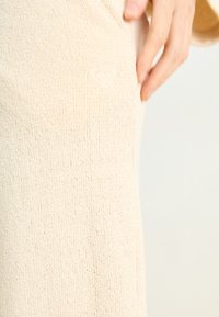 Beige strikket topp med teksturert stoff og et preget hjerte-logo på siden. Enkel design med en avslappet passform.