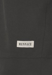 Tissu noir à texture lisse avec une étiquette blanche cousue, affichant le mot "MENNACE" en lettres majuscules noires.