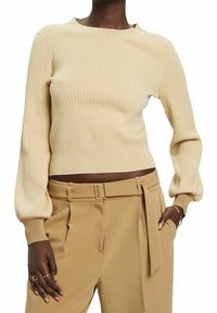 Pull beige côtelé à manches longues bouffantes et large encolure, associé à un pantalon beige sur mesure avec ceinture et poches latérales.
