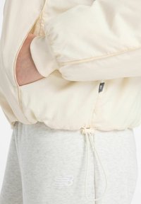 Lichtbeige bomberjack met ribbelmanchetten en zijzak, gecombineerd met lichtgrijze joggingsbroek met geborduurd logo en verstelbaar trekkoord.