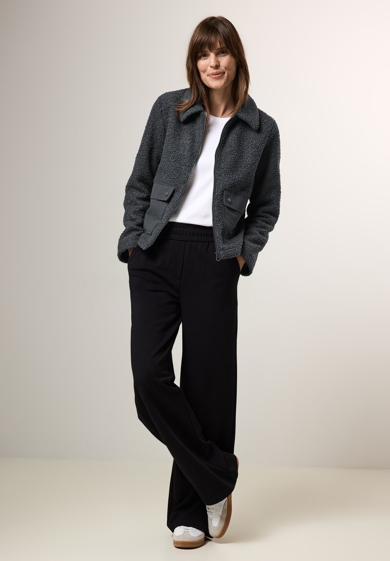 Veste en polaire gris texturé avec fermeture éclair et poches à rabat, associée à un pantalon noir à jambes larges et des baskets blanches. Look épuré et décontracté.