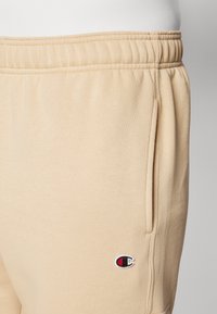 Béžové tepláky s elastickým pásom, so bočnými vreckami a malým vyšitou logom v červenej a námorníckej farbe na ľavom stehne.