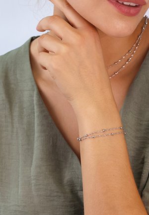 Silver chain bracelet with small spherical accents, worn on wrist. Model's hand close to face, wearing a green textured top.