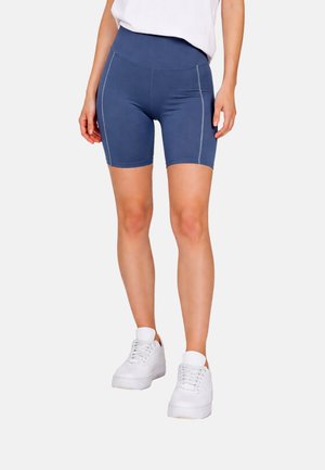 Frau trägt dunkelblaue, eng anliegende Shorts auf Oberschenkellänge mit hohem Bund, weiße Sneakers und ein weißes Top, steht vor einem einfarbigen Hintergrund.