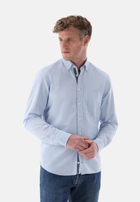 Hellblaues Button-Down-Hemd mit Kragen, einer einzelnen Brusttasche und kontrastierenden Innendetails. Der Stoff wirkt glatt und leicht strukturiert.