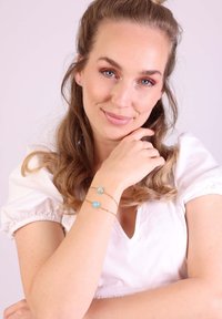 Gouden kettingarmband met twee turquoisekleurige stenen. Het model draagt een witte top met pofmouwen en heeft golvend haar. De huid ziet er glad uit.