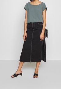 Top grigio a maniche corte abbinato a una gonna midi in denim nera con cuciture bianche e spacco laterale, completato da sandali neri.