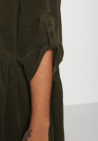 Bras avec un petit tatouage partiellement dissimulé par une manche en velours côtelé vert olive remontée avec une patte de bouton sur fond uni.