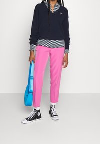 Pull marin bleu avec un col en V, chemise à col à motifs en dessous, pantalon rose tapered, baskets montantes noires et sac à bandoulière turquoise.