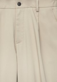 Gros plan sur un pantalon beige montrant le bouton frontal, les passants de ceinture et les coutures de la poche latérale sur un tissu texturé.