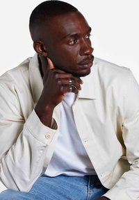 Witte casual jas met knoopmanchetten, gedragen over een wit t-shirt. De achtergrond is eenvoudig. Handen rusten op de kin, met zichtbare metalen ringen.