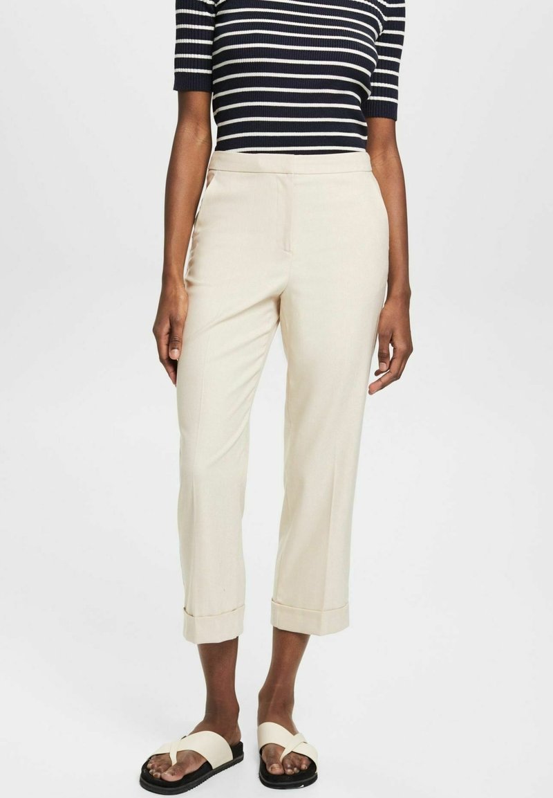 Pantalon beige court ajusté, avec une texture lisse et des revers roulés. Porté avec un haut à manches courtes rayé bleu marine et des sandales.