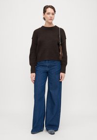 Pulover tricotat maro cu umeri căzuți, asortat cu blugi din denim albastru cu croială largă. Include o curea cu lanț și detaliu cu franjuri.