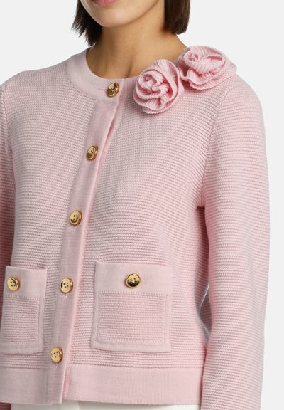 Svetlo roza kardigan z teksturiranim tkanjem, ki ima dve sprednji žepi, zlatimi gumbi in dvema rožnima detajlama iz blaga na rami.
