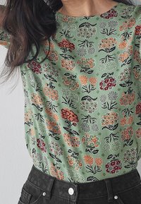Top floral verde con mangas cortas, que presenta patrones de flores rojas, naranjas y negras sobre una tela texturizada. Combinado con pantalones negros de talle alto.
