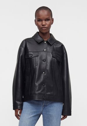 Personne debout face à l’avant, portant une veste noire en cuir à boutons, avec col et poches poitrine, et un jean bleu clair sur un fond uni.