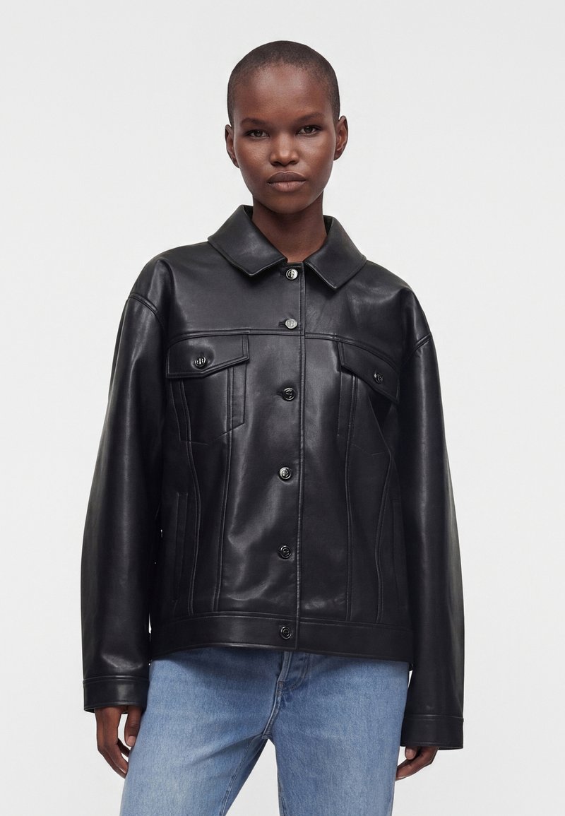 Personne debout face à l’avant, portant une veste noire en cuir à boutons, avec col et poches poitrine, et un jean bleu clair sur un fond uni.