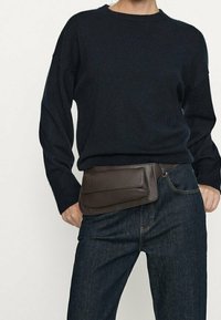 Mörkbrun läder midjeväska med två platta fickor, bärs över mörk denimjeans; ihop med en svart stickad tröja. Enkel, funktionell design.