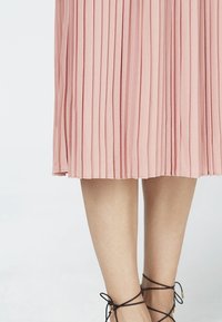 Rosa Plissee-Rock in mittellanger Form, getragen mit schwarzen Schnürsandalen, die die Unterschenkel und Füße vor einem weißen Hintergrund zeigen.