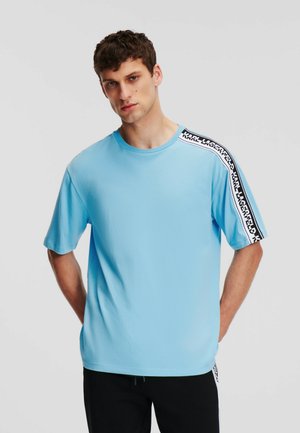 Light blue cotton t-shirt with short sleeves and a round neckline, featuring a contrasting black logo band along one shoulder.