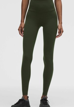 Gambe con leggings verde scuro a vita alta e scarpe da ginnastica nere, in piedi davanti a uno sfondo chiaro uniforme.