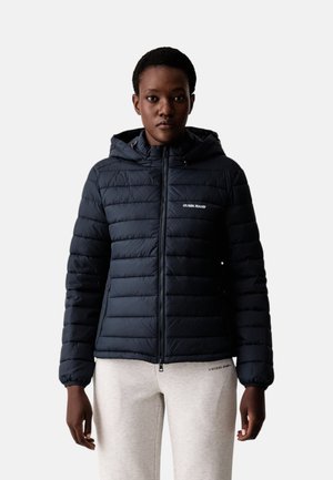 Persona che indossa una giacca nera trapuntata con cappuccio e zip e pantaloni bianchi, in piedi davanti a uno sfondo chiaro e uniforme.