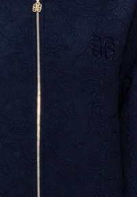 Granatowa kurtka na zamek błyskawiczny z teksturowanymi wzorami fal, wyposażona w złoty metalowy zamek i haftowane logo na piersi. Miękki materiał.