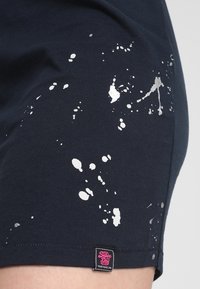 Pantaloni corti in cotone blu navy con un design a macchie di vernice argentata, con un'etichetta del marchio ben visibile all'orlo. Tessuto morbido e forma aderente.