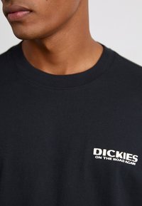 T-shirt in cotone nero con collo rotondo e maniche corte. Il testo stampato in bianco sul lato superiore sinistro recita "DICKIES ON THE ROAD AGAIN."