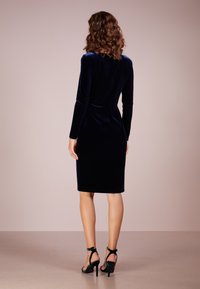 Žena s kudrnatými hnědými vlasy, která má na sobě přiléhavé, dlouhé námořnické sametové šaty ke kolenům a ladící vysoké podpatky, pohled zepředu.