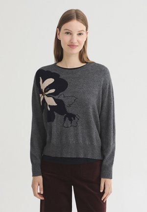 Jeune femme aux cheveux bruns et raides porte un pull gris foncé avec un grand motif floral noir et beige sur le côté gauche, et un pantalon foncé.