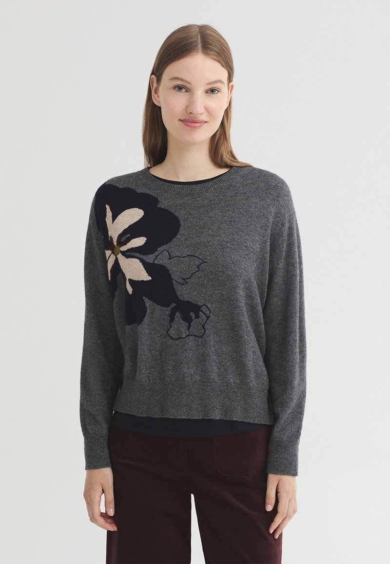 Jeune femme aux cheveux bruns et raides porte un pull gris foncé avec un grand motif floral noir et beige sur le côté gauche, et un pantalon foncé.