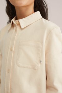 Gai smilšu krāsas korduroja krekls ar nolaižamu apkakli, priekšējo pogu aizdari un krūšpocketu, kurā ir neliels sirds birkas. Jauns