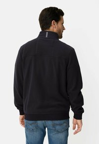 Dunkelblaue Fleecejacke mit hohem Kragen, gerippten Bündchen und einer dezenten Nahtverzierung am Rücken. Kombiniert mit blauen Jeans.