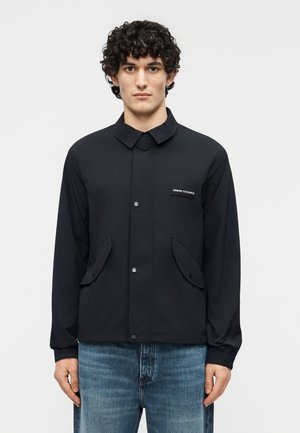 Lokkis juustega mees, kes kannab musta nööpidega jopet, millel on kaks esimest taskut ja Armani Exchange'i logo, kokku sobitatud siniste teksapükstega.